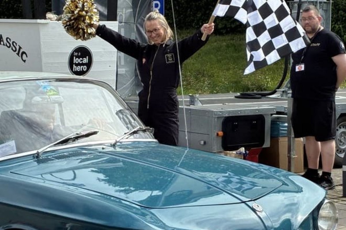 Eine strahlende Frau mit einer Zielflagge und einem Blumestrauß, sie bejubelt ein Auto
